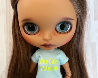 Op de afbeelding: Een pop met bruin haar en grote blauwe ogen. De pop draagt een lichtblauw t-shirt met de tekst "One Cute Chick" in geel en een kleine gele kuikengrafiek. De pop draagt ook een blauwe spijkerbroek.