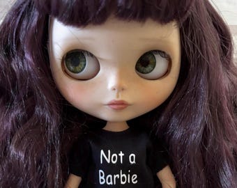 Op de afbeelding: Een pop met lang, donkerpaars haar en grote groene ogen. De pop draagt een zwart t-shirt met de witte tekst "Not a Barbie". De pop heeft een bleke teint met roze wangen en een neutrale uitdrukking.