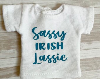 Op de afbeelding: Een klein wit T-shirt met de tekst "Sassy IRISH Lassie" in turquoise letters. Het shirt heeft korte mouwen en een ronde halslijn. De stof lijkt een gebreid materiaal te zijn.