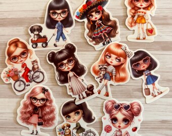 Op de afbeelding: Een verzameling uitgestanste stickers met gestileerde illustraties van poppen. De poppen hebben grote ogen, brillen en verschillende kapsels. Sommige poppen worden afgebeeld met huisdieren, fietsen en accessoires, in verschillende outfits en poses.