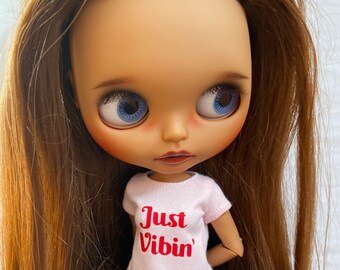 Op de afbeelding: Een pop met lichtbruin haar en blauwe ogen. De pop draagt een roze t-shirt met de woorden "Just Vibin'" in het rood. De pop draagt ook een blauwe spijkerbroek. De pop heeft een lichte huidskleur en roze wangen.
