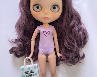 Op de afbeelding: Een pop met lang, golvend paars haar, die een paarse badpak met een hartvormig ontwerp draagt. De pop houdt een witte draagtas met blauwe stippen en de tekst "Malibu Beach".