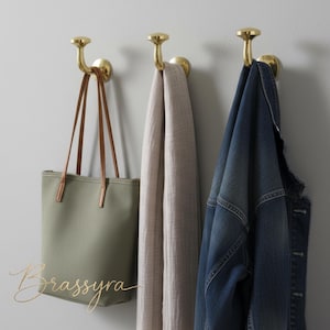 May include: Three brass wall hooks hold a green tote bag with brown straps, a light beige scarf, and a blue denim jacket. The hooks are mounted on a light gray wall, offering a stylish storage solution.