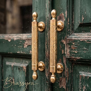 May include: Two vertical brass door handles with intricate engraved patterns, featuring spherical finials at the top and bottom. The handles are mounted on a dark green wooden door with visible paint chipping and wear, showing a metallic gold finish.