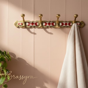 May include: A brass wall-mounted coat rack with four hooks. The rack has red and white diamond-shaped detailing. A cream towel hangs from one of the hooks. The word "Brassyra" is written in cursive on the lower left.