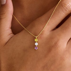 May include: A gold chain necklace featuring a vertical pendant with three small, round, faceted gemstones arranged in a row. The top stone is yellow, the middle stone is clear white, and the bottom stone is a vibrant pink, all set in gold.