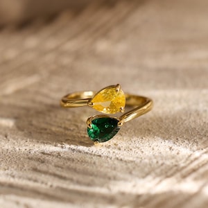 May include: A gold ring with a distinctive design, showcasing two pear-cut gemstones. One gemstone is a vibrant yellow, the other a deep green. The ring's band curves to cradle the stones. The ring is displayed on a textured surface, with soft lighting.