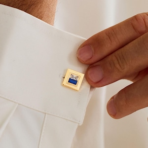 May include: Gold square cufflinks with a blue and white design. The cufflinks are on a white dress shirt. The image shows a person adjusting the cufflink.