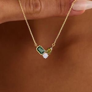Könnte beinhalten: Eine goldene Halskette mit einem Anhänger, der aus einem rechteckigen smaragdgrünen Stein, einem birnenförmigen olivgrünen Stein und einem kleinen runden klaren Stein besteht. Die zarte Goldkette liegt am Hals und betont die Anordnung der Edelsteine.