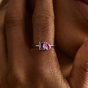 Peut inclure: Une bague en argent ornée de deux pierres roses. L'une est rectangulaire, l'autre en forme de poire. La bague est portée sur un doigt, mettant en valeur le design et la couleur du bijou.