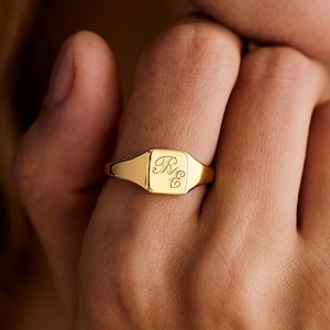 May include: A gold signet ring with engraved initials. The ring features a rectangular face with rounded corners and a polished finish. The initials "R C" are elegantly inscribed on the face. The ring is shown on a finger.