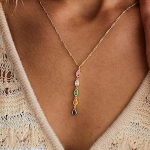 Puede incluir: Un collar dorado con un colgante con una serie de piedras preciosas de colores en forma de lágrima. Las piedras preciosas incluyen rosa, blanco, verde y azul. El collar se lleva sobre una parte superior clara.