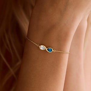 May include: A delicate gold bracelet featuring two teardrop-shaped gemstones, one clear and one blue. The bracelet is connected by a fine chain and is worn on a wrist. The overall design is elegant and minimalist.