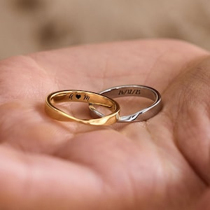 May include: Two wedding rings, one gold and one silver, rest in an open palm. The gold ring has a heart symbol and the initials "A & M" engraved. The silver ring is engraved with "26/12/15".