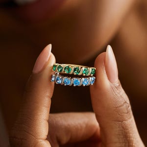 May include: Two gold rings, each with a row of gemstones. One ring features green gemstones, the other blue. The rings are held up by a person with a blurred background.