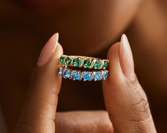 Pode incluir: Dois anéis de ouro, cada um com uma fileira de pedras preciosas. Um anel apresenta pedras verdes, o outro azuis. Os anéis são segurados por uma pessoa com um fundo desfocado.