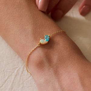 Peut inclure: Un bracelet à fine chaîne dorée orné de deux petites pierres précieuses en forme de goutte, serties dans des montures dorées. L'une est une opale blanche laiteuse et irisée, l'autre une topaze bleue translucide, placées côte à côte.