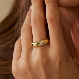 May include: A gold band ring featuring a small, round, olive-green gemstone set next to a tiny, rectangular, clear white stone. The ring has a smooth, polished finish and a rounded profile, showcasing a minimalist design with two distinct stones.