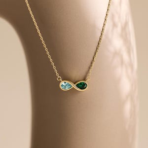 May include: A gold chain necklace featuring an infinity symbol pendant. The pendant is set with a light blue pear-shaped gemstone on one side and a dark green pear-shaped gemstone on the other side, connected by a thin gold metal frame.