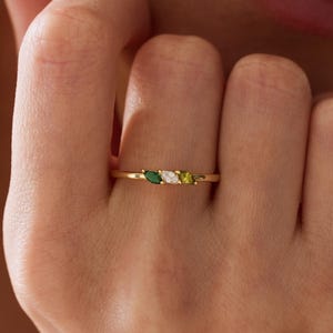 May include: A gold ring featuring three marquise-cut gemstones arranged horizontally. The stones consist of a dark green gem on the left, a clear white gem in the center, and a light olive green gem on the right, all set on a thin gold band.