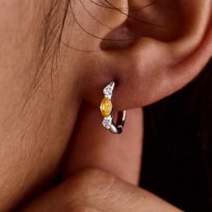 May include: A silver hoop earring featuring a yellow oval gemstone and two smaller white gemstones. The earring is displayed on an ear, highlighting the jewellery's design. The gemstones are set in silver metal.