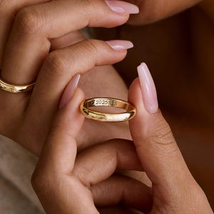 May include: Two gold wedding bands are held in a person's hands. One ring is on a finger, and the other is being held up to the camera. The ring held up has the inscription "20251125" engraved on it. The rings are shiny and reflect the light.