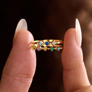 May include: A gold ring featuring two thin bands connected by a series of small, round, multi-coloured gemstones including blue, yellow, pink, green, and clear stones set in a delicate, beaded metal design that creates a layered appearance.