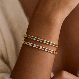 May include: Two gold bangles are displayed on an arm. One is adorned with green gemstones, the other with blue. The jewellery is set against a soft, neutral background, highlighting the bracelets' design and colour.