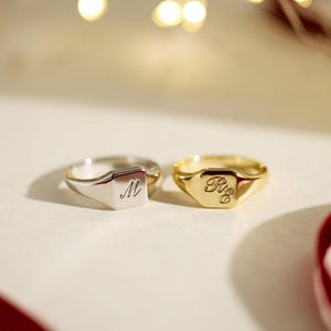 May include: Two signet rings, one silver and one gold, are displayed. The silver ring features the letter "M" engraved on a square face. The gold ring has the initials "R&E" engraved on its face. Both rings are on a white surface.