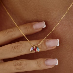 Könnte beinhalten: Eine goldene Kettenkette mit drei kleinen Anhänger-Anhängern. Jeder Anhänger ist blattförmig und mit einem andersfarbigen Edelstein besetzt: lila, rot und blau.