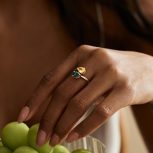May include: A gold ring with two pear-cut gemstones, one yellow and one green. The ring is on a hand with manicured nails. A bowl of green grapes is in the background.