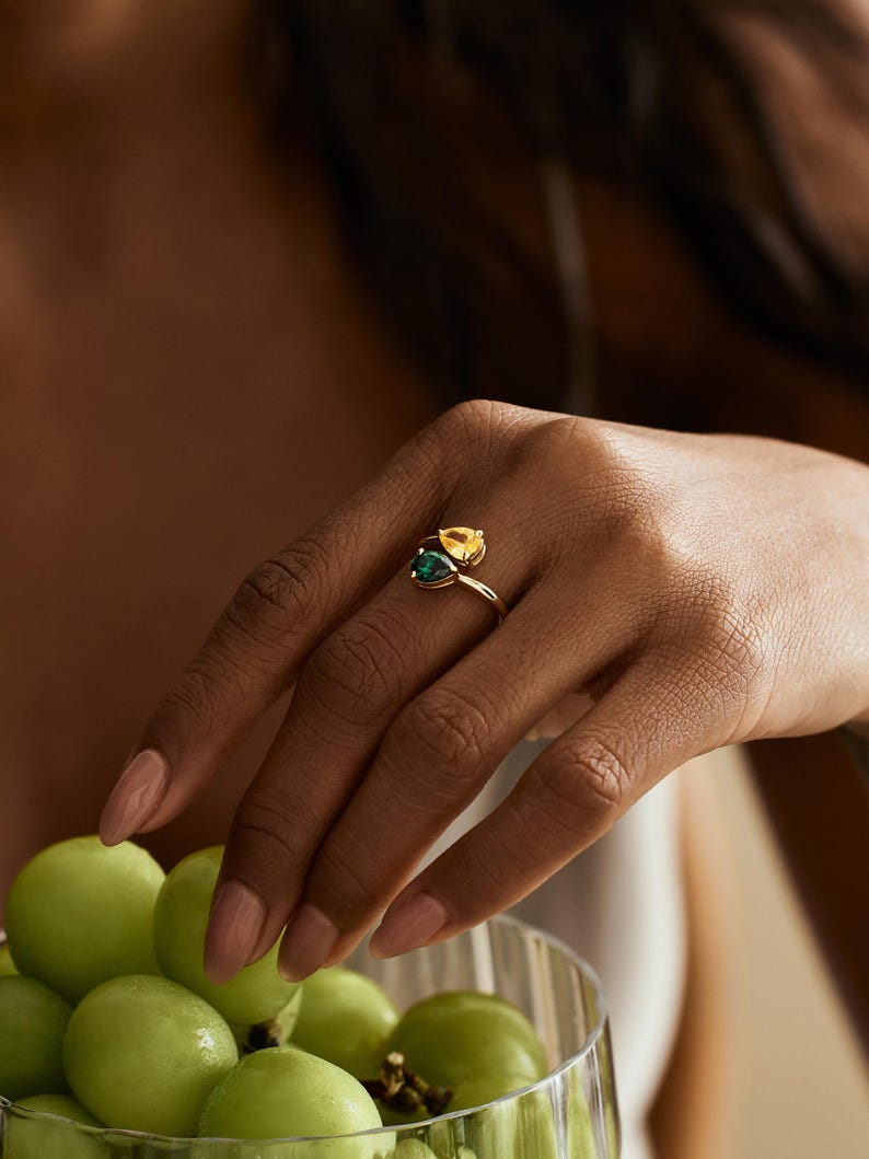 May include: A gold ring with two pear-cut gemstones, one yellow and one green. The ring is on a hand with manicured nails. A bowl of green grapes is in the background.