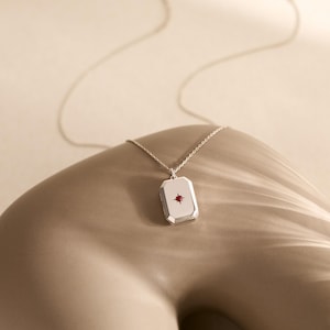 Puede incluir: Un collar con colgante rectangular plateado con una peque&ntilde;a gema roja en el centro. El colgante cuelga de una delicada cadena plateada. El collar se exhibe sobre una superficie de tono neutro.