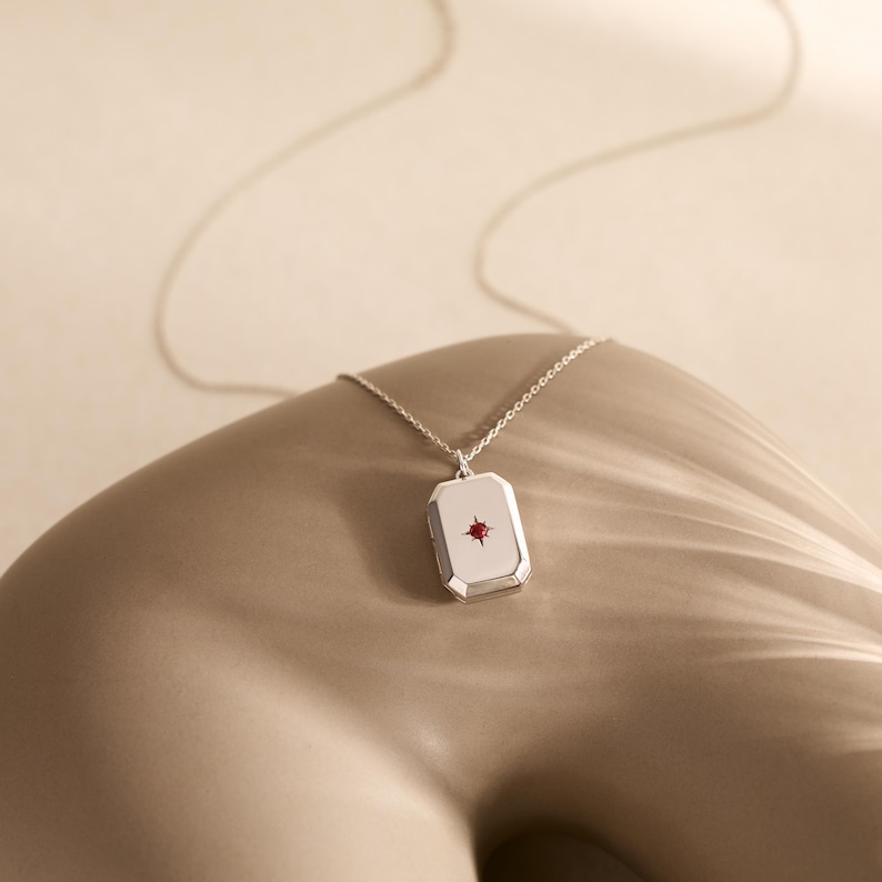 Puede incluir: Un collar con colgante rectangular plateado con una peque&ntilde;a gema roja en el centro. El colgante cuelga de una delicada cadena plateada. El collar se exhibe sobre una superficie de tono neutro.