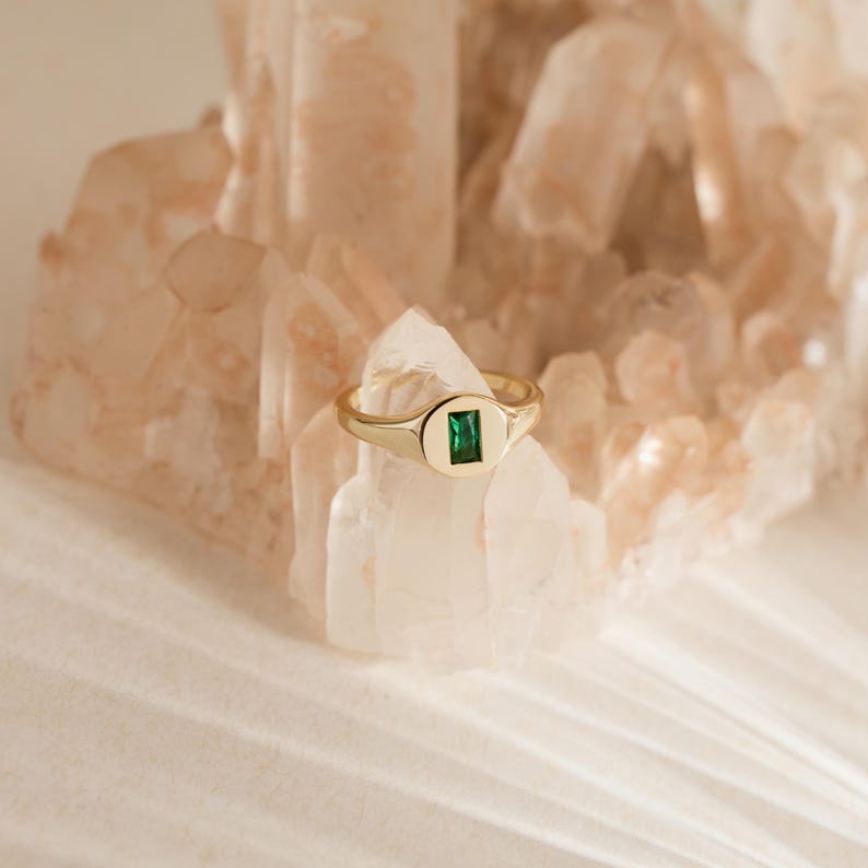 May include: A gold ring with a rectangular emerald-green stone set in a smooth, oval-shaped bezel. The ring is displayed on a light-colored, crystalline surface, creating a contrast between the warm gold and the cool green gemstone.