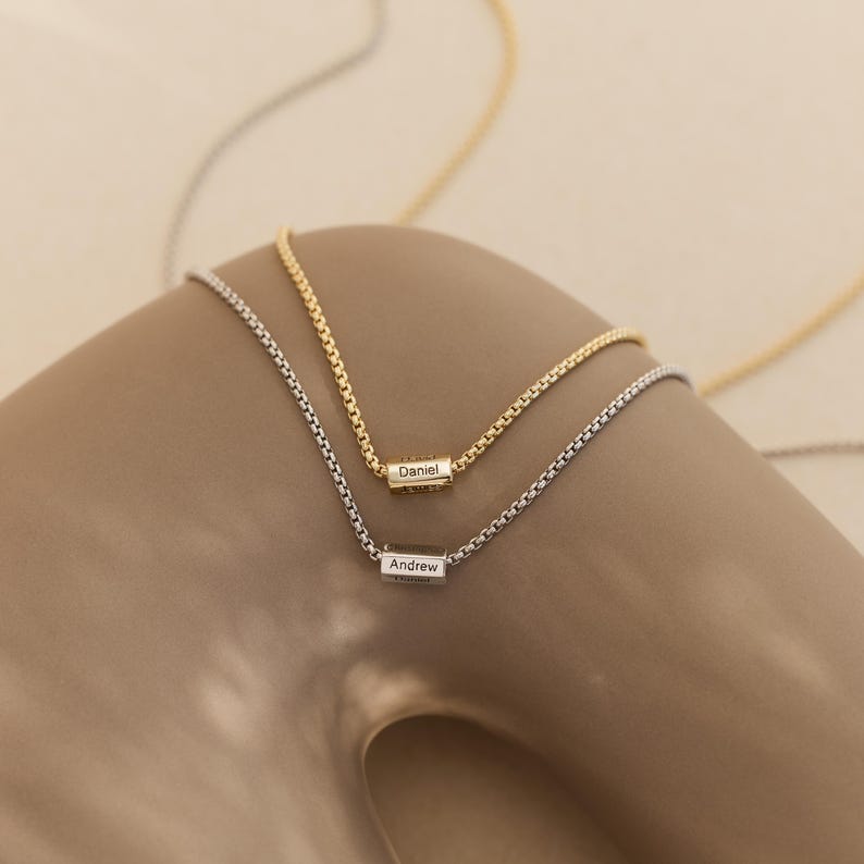 May include: Two delicate gold and silver necklaces with personalized name tags. The gold necklace has the name "Daniel" engraved on the tag, and the silver necklace has the name "Andrew" engraved on the tag.