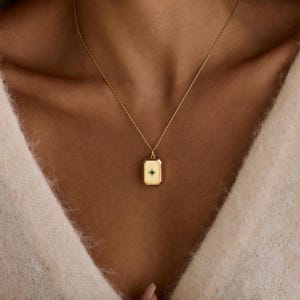 Puede incluir: Un collar con colgante rectangular de color dorado con una peque&ntilde;a piedra preciosa verde en el centro. El colgante cuelga de una delicada cadena dorada. El collar se lleva sobre un su&eacute;ter color crema.