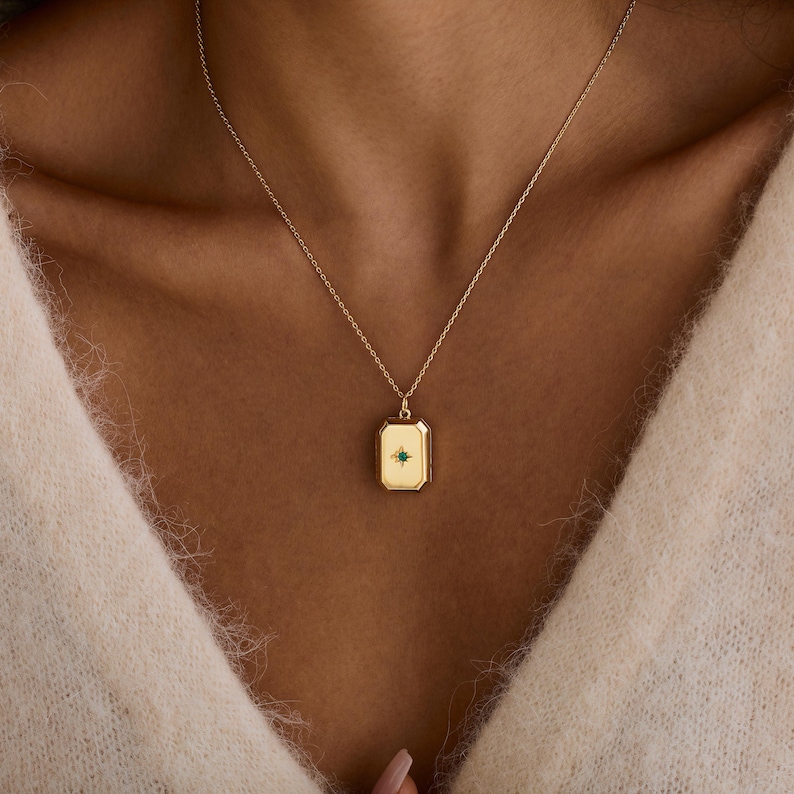 Puede incluir: Un collar con colgante rectangular de color dorado con una peque&ntilde;a piedra preciosa verde en el centro. El colgante cuelga de una delicada cadena dorada. El collar se lleva sobre un su&eacute;ter color crema.
