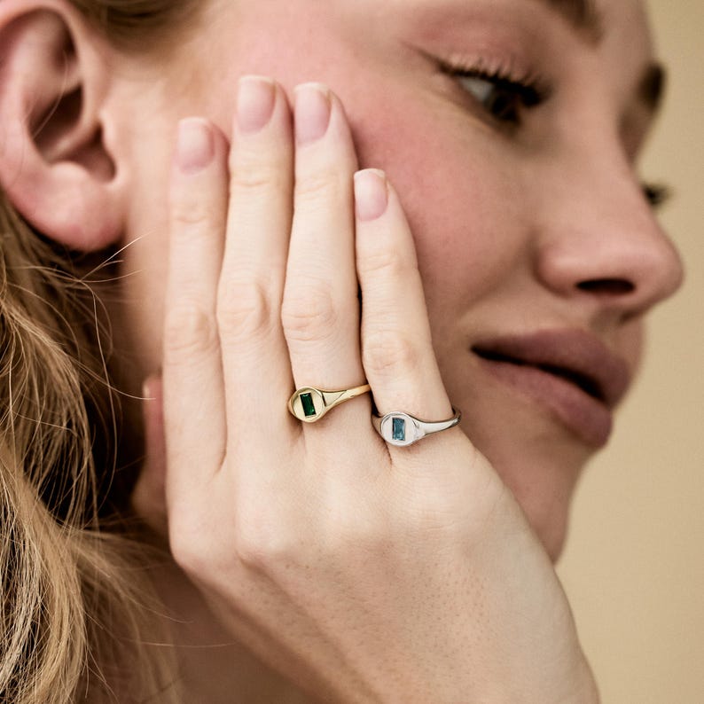 May include: Two signet rings, one gold and one silver, each featuring a rectangular gemstone. The gold ring has a green stone, while the silver ring has a blue stone. The rings are on a hand, with the background out of focus.