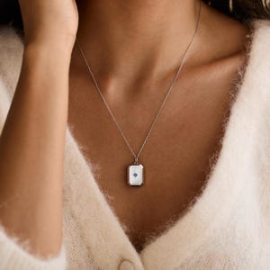 Puede incluir: Un collar plateado con un colgante rectangular. El colgante tiene un fondo blanco con un peque&ntilde;o dise&ntilde;o de estrella azul. El collar se lleva sobre un su&eacute;ter color crema.