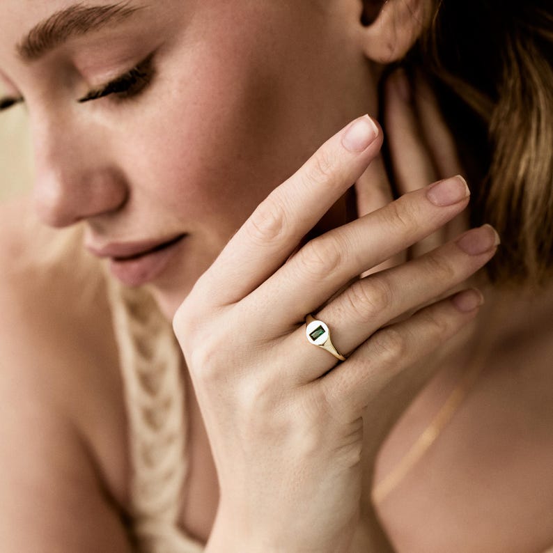 May include: A gold ring with a rectangular green gemstone. The ring is on a finger. The ring has a simple, elegant design.