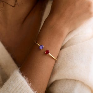 May include: A gold-colored bracelet with a red and a blue oval-shaped gemstone. The bracelet is open-ended and worn on the wrist. The gemstones are set on either end of the bracelet.