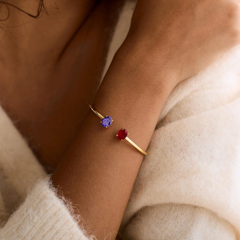 May include: A gold-colored bracelet with a red and a blue oval-shaped gemstone. The bracelet is open-ended and worn on the wrist. The gemstones are set on either end of the bracelet.