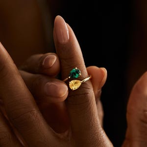 May include: A gold ring with a pear-shaped yellow gemstone and a round green gemstone. The ring is held up by a person's fingers, against a dark background.