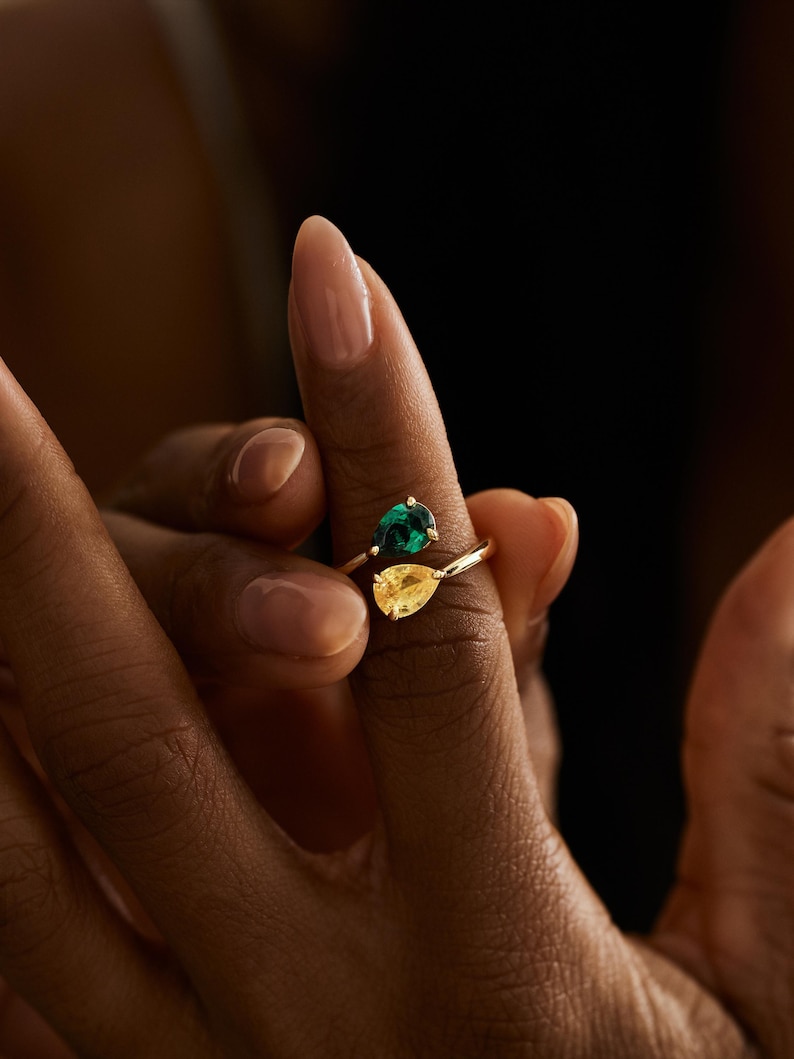 May include: A gold ring with a pear-shaped yellow gemstone and a round green gemstone. The ring is held up by a person's fingers, against a dark background.