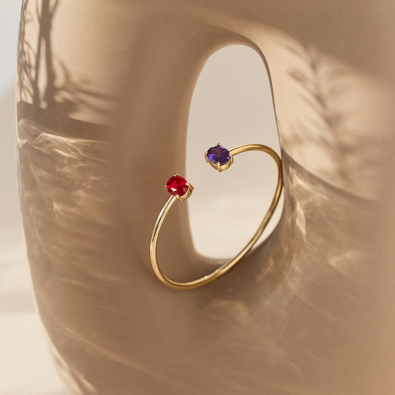 May include: A gold-colored open cuff bracelet with a red and purple gemstone at each end. The bracelet is set against a neutral background, with shadows adding depth. The gemstones are round and faceted, catching the light.
