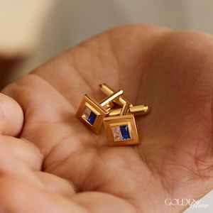Könnte beinhalten: Ein Paar goldfarbene Manschettenknöpfe mit quadratischen Flächen. Jeder Manschettenknopf hat ein rechteckiges blaues und weißes Edelsteindesign. Die Manschettenknöpfe werden in einer offenen Hand gehalten.