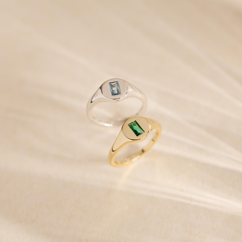 May include: Two signet rings, one silver and one gold, each featuring a rectangular gemstone. The silver ring has a light blue stone, while the gold ring showcases a green stone. Both rings are set against a soft, neutral background.