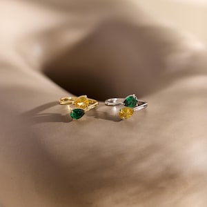 May include: Two rings, one gold and one silver, each with a pair of pear-cut gemstones. The gold ring has yellow and green stones, the silver ring has green and yellow stones. The rings are displayed against a neutral backdrop.