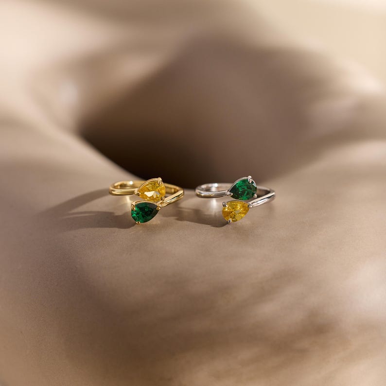 May include: Two rings, one gold and one silver, each with a pair of pear-cut gemstones. The gold ring has yellow and green stones, the silver ring has green and yellow stones. The rings are displayed against a neutral backdrop.